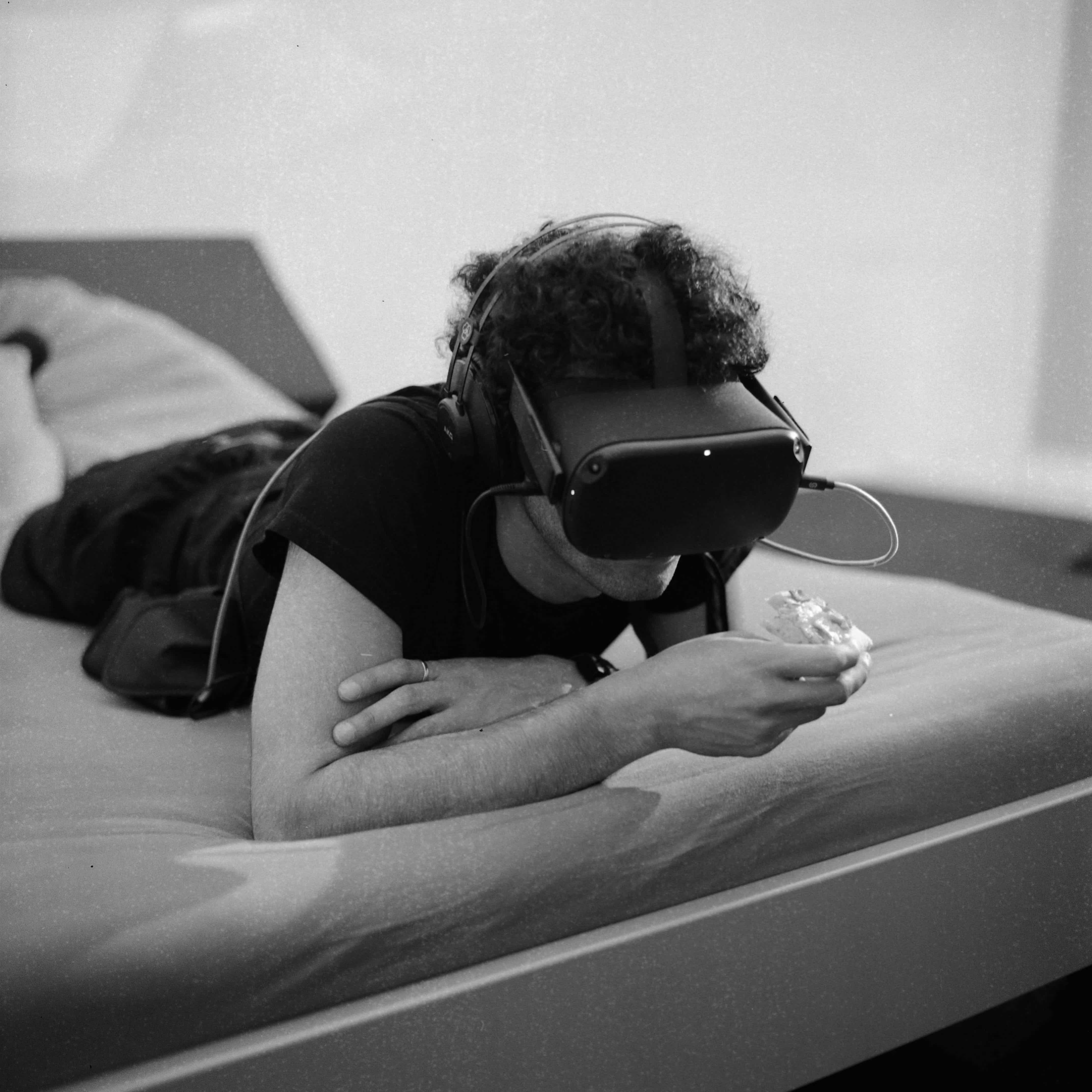 The artist with the VR headset on, eating pizza in bed.